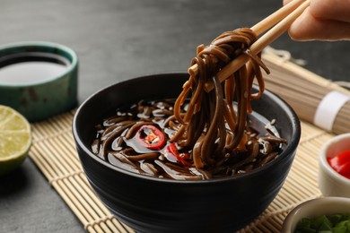 Photo of Woman eating delicious buckwheat noodle (soba) soup with chopsticks at grey table, closeup