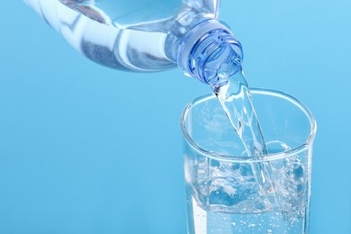 Pouring water from bottle into glass on light blue background, closeup