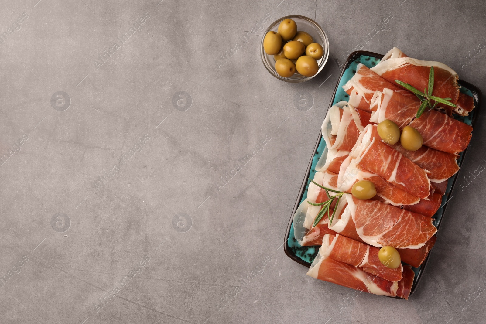 Photo of Rolled slices of delicious jamon with rosemary and olives on grey table, flat lay. Space for text