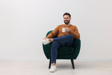 Photo of Handsome man with cup of drink sitting in armchair near white wall indoors