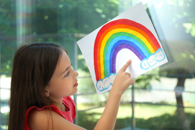 Photo of Little girl with picture of rainbow near window indoors.  Stay at home concept