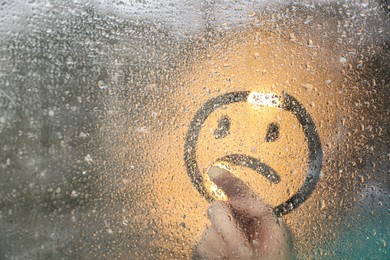 Woman drawing sad face on foggy window at rainy weather