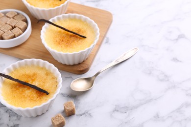 Delicious creme brulee in bowls, vanilla pods, sugar cubes and spoon on white marble table, space for text