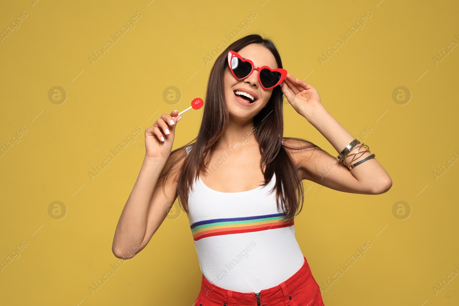 Photo of Portrait of beautiful young woman with heart shaped sunglasses and lollipop on color background