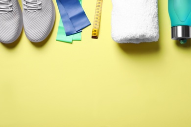 Photo of Sneakers, fitness elastic band, measuring tape, towel and bottle of water on yellow background, flat lay. Space for text
