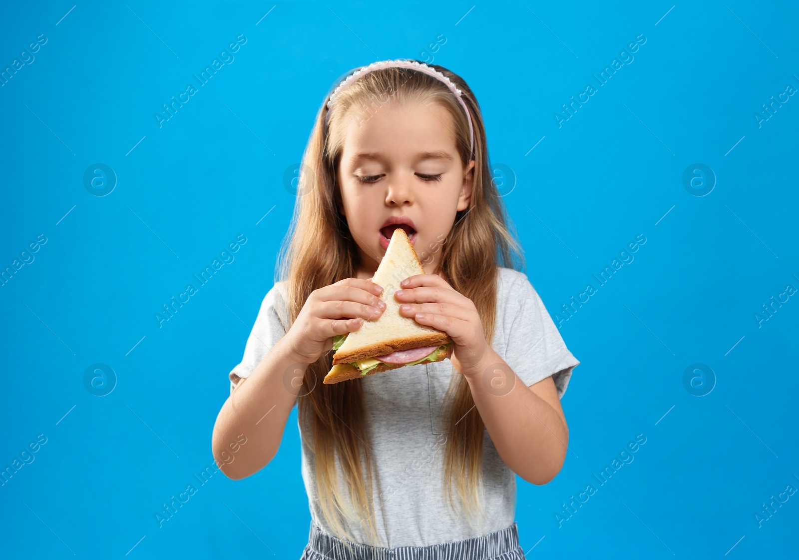 Photo of Cute little girl eating tasty sandwich on light blue background