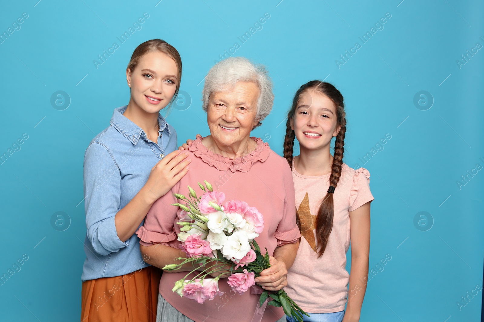 Photo of Happy sisters with their grandmother holding flowers on light blue background