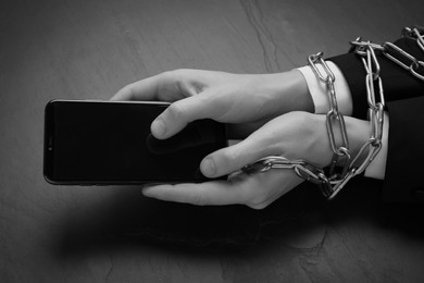 Closeup view of internet addicted woman with chained hands using smartphone at dark table, space for text. Black and white effect