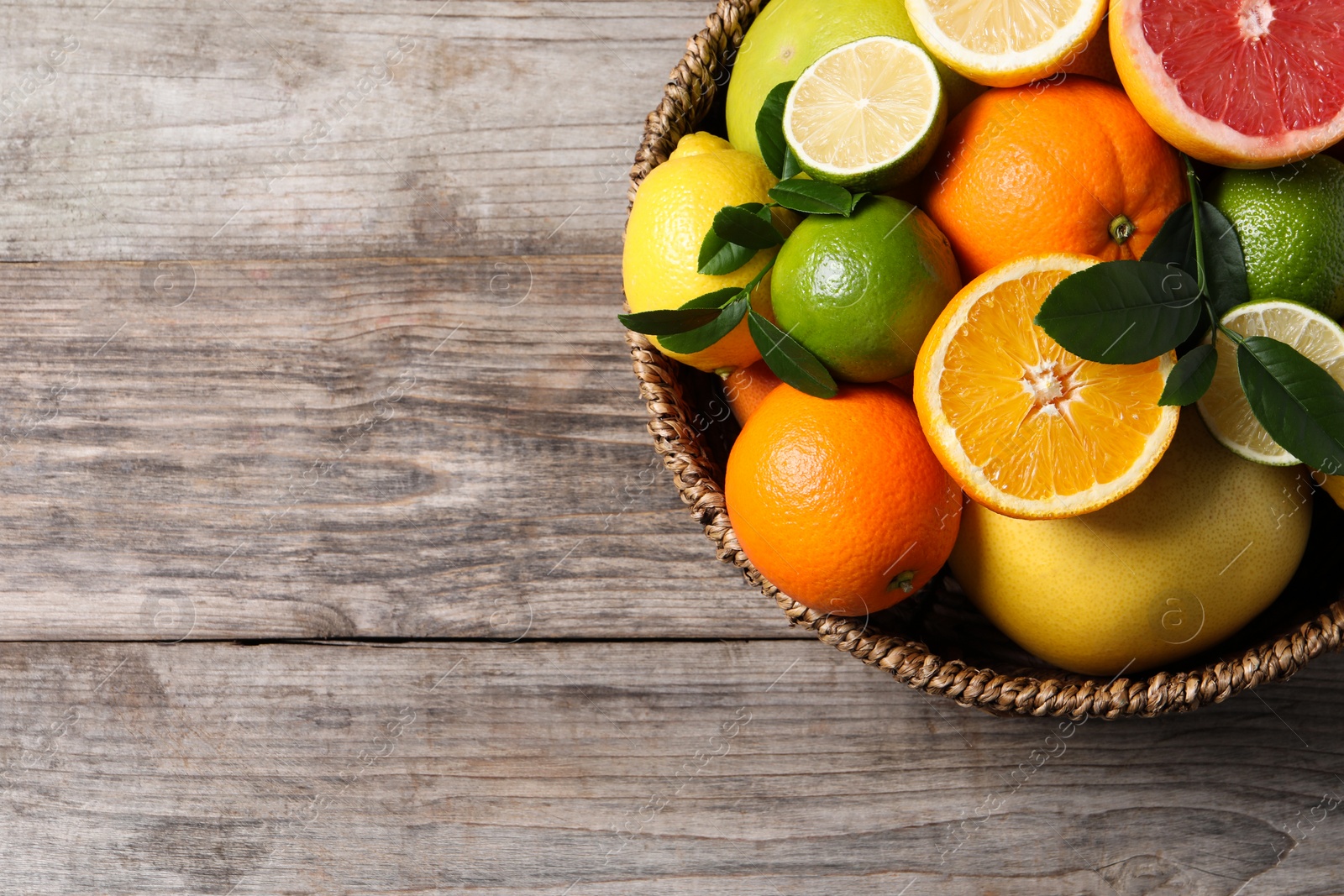 Photo of Different fresh citrus fruits and leaves in wicker basket on wooden table, top view. Space for text