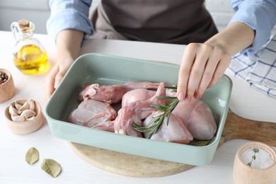 Woman putting rosemary into baking dish with raw rabbit meat at white wooden table, closeup