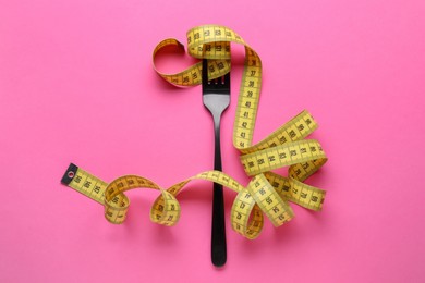 Fork with measuring tape on pink background, top view. Diet concept