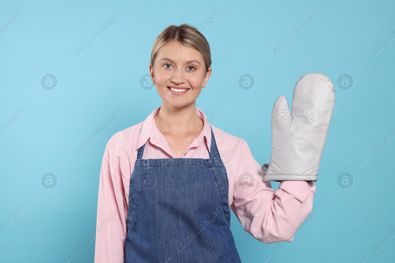 Photo of Beautiful young woman in denim apron and oven glove on light blue background