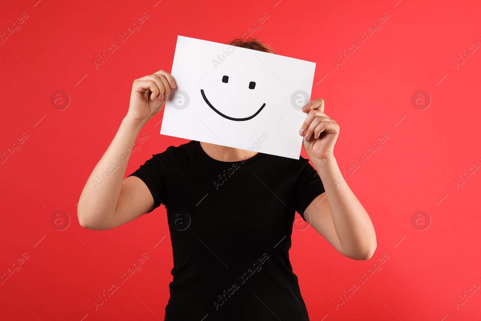 Photo of Woman hiding behind sheet of paper with happy face on red background