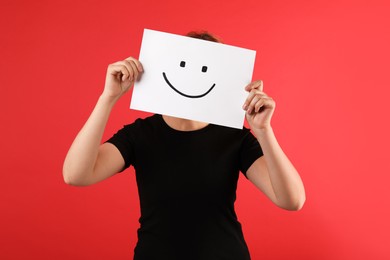 Woman hiding behind sheet of paper with happy face on red background