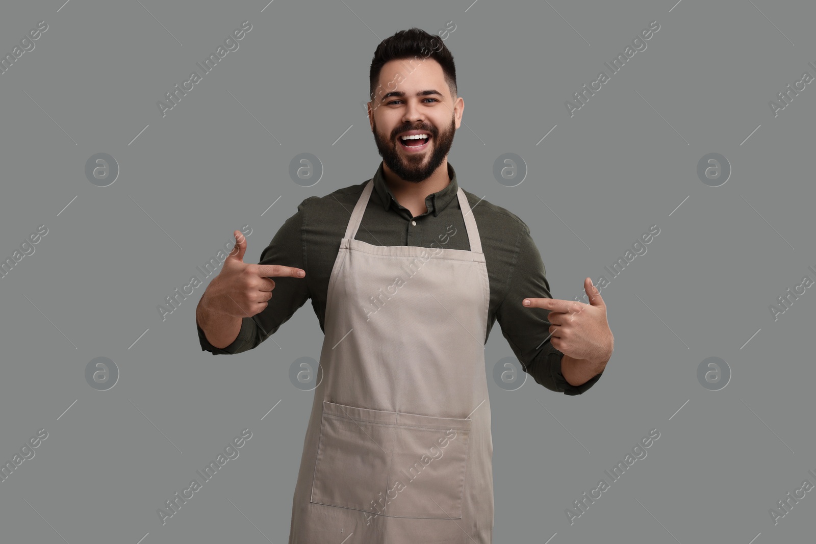 Photo of Happy man pointing at kitchen apron on grey background. Mockup for design