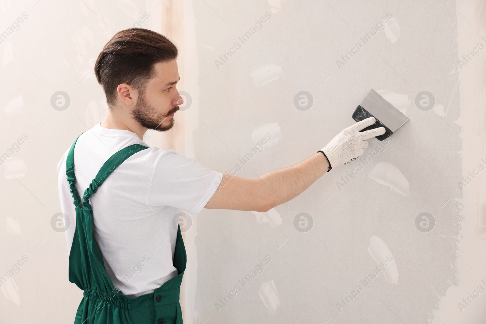 Photo of Worker in uniform plastering wall with putty knife indoors