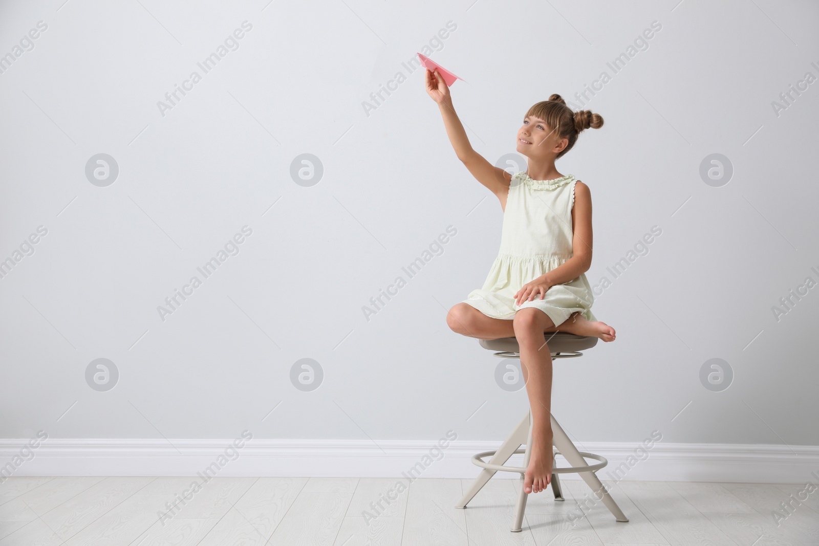 Photo of Cute little girl playing with paper plane near light grey wall. Space for text