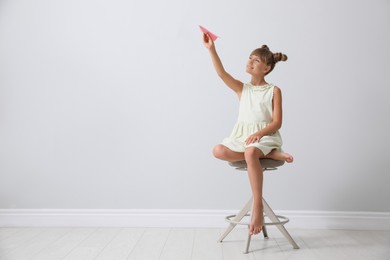 Cute little girl playing with paper plane near light grey wall. Space for text