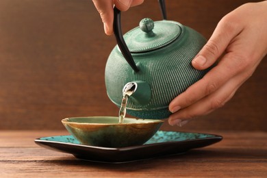 Photo of Woman pouring freshly brewed tea from teapot into cup at wooden table, closeup. Traditional ceremony
