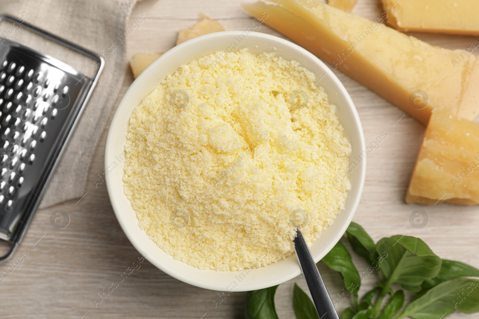 Photo of Delicious grated parmesan cheese in bowl, grater and basil on white wooden table, flat lay