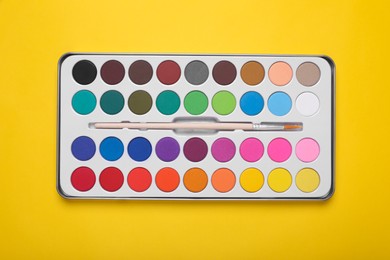 Photo of Watercolor palette with brush on yellow background, top view