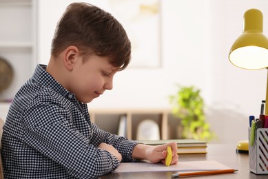 Photo of Little boy erasing mistake in his notebook at wooden desk indoors