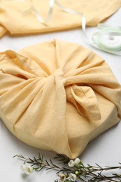 Photo of Furoshiki. Things packed in fabric, ribbon and flowers on white table, closeup