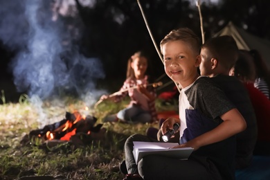 Photo of Little boy with book and flashlight near bonfire at night. Summer camp