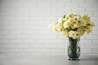 Beautiful white Eustoma flowers in vase on table near brick wall. Space for text