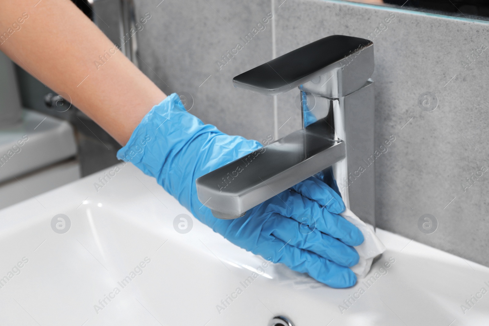 Photo of Woman in glove cleaning faucet of bathroom sink with paper towel, closeup