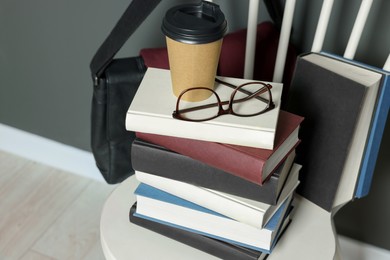 Photo of Stack of different books, paper cup and glasses on white chair indoors