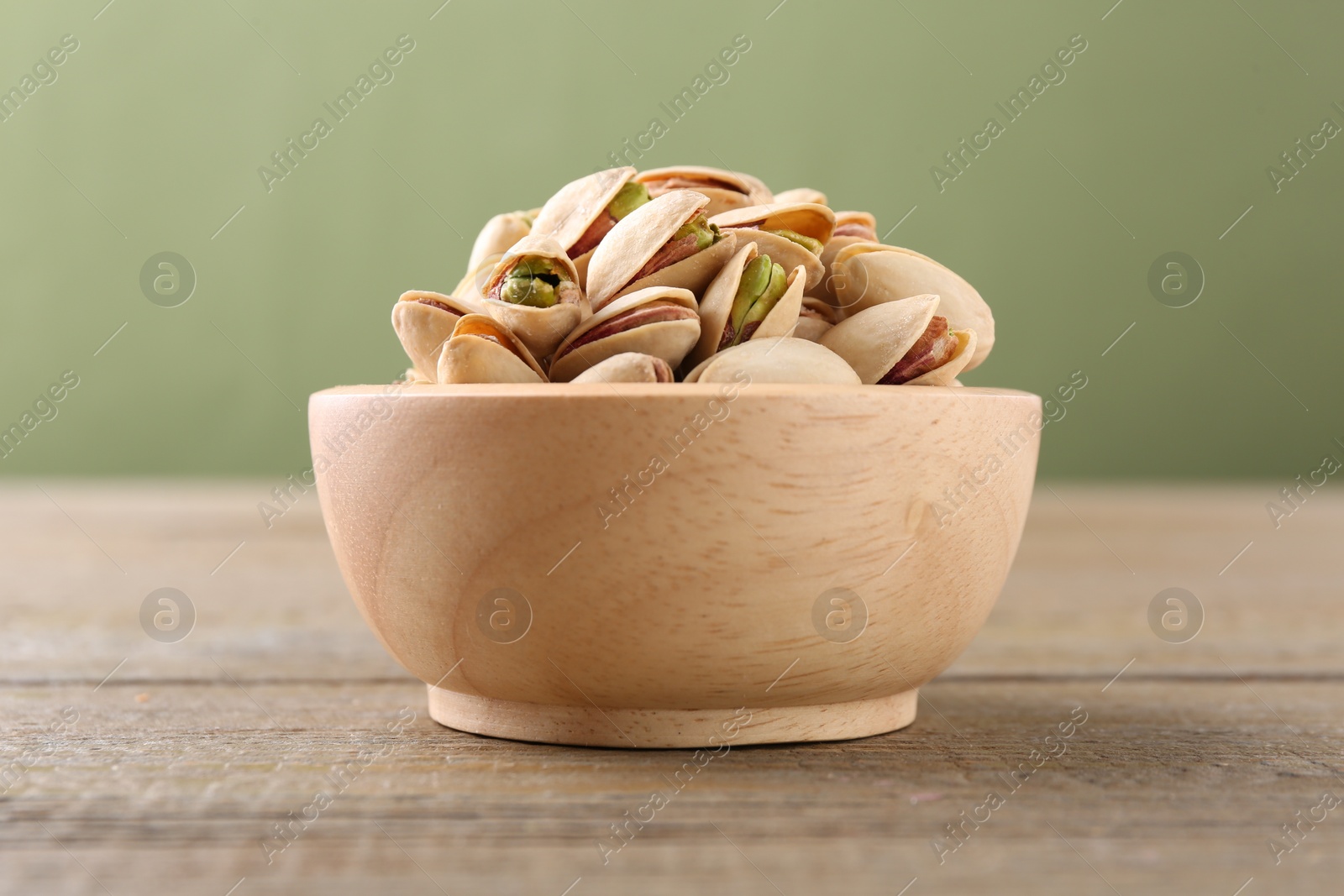 Photo of Tasty pistachios in bowl on wooden table against olive background, closeup