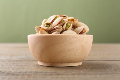 Tasty pistachios in bowl on wooden table against olive background, closeup