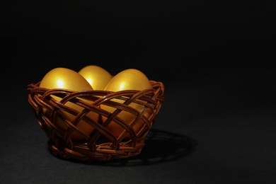 Photo of Shiny golden eggs in wicker bowl on black background, space for text