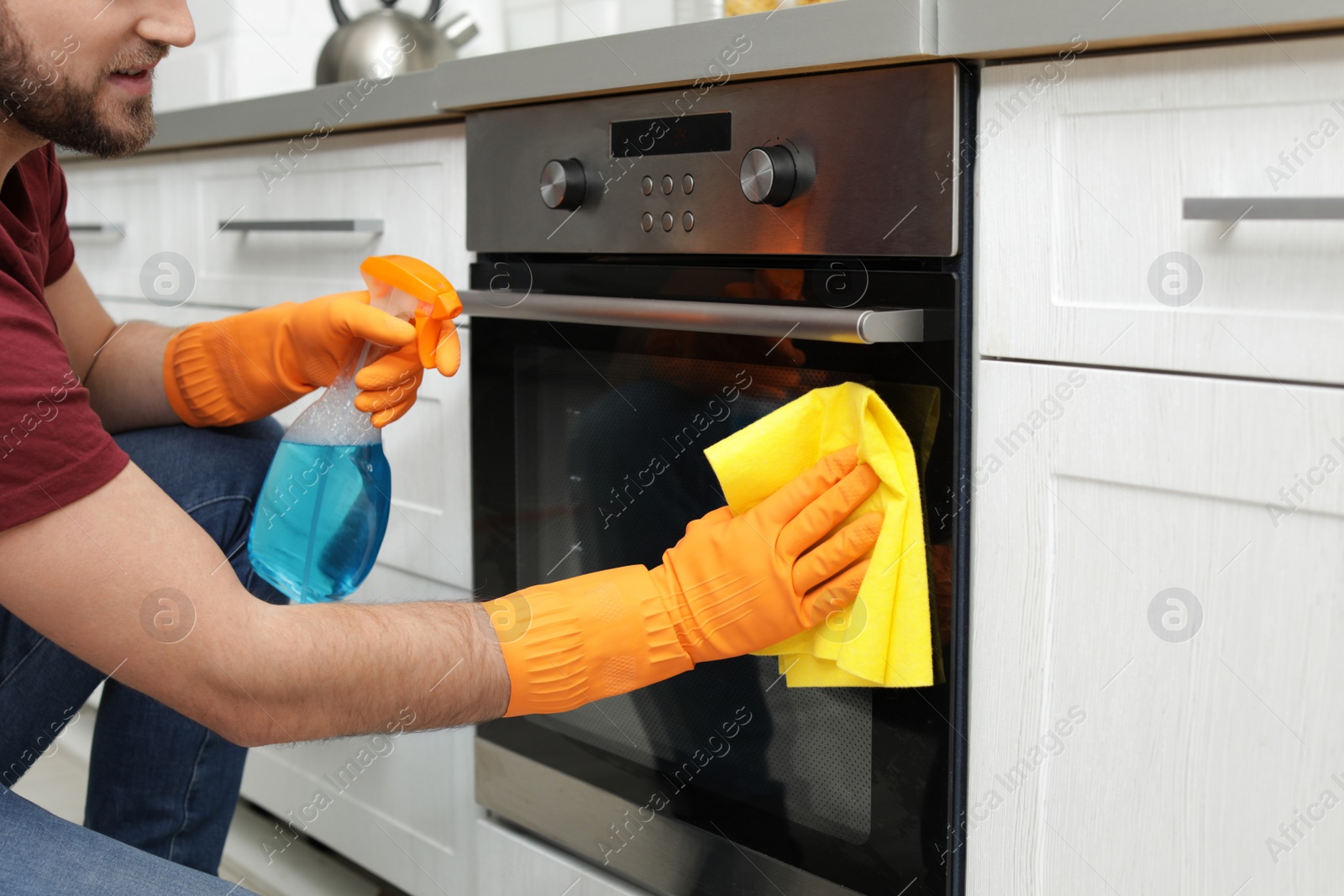 Photo of Young man cleaning oven with rag and detergent in kitchen, closeup