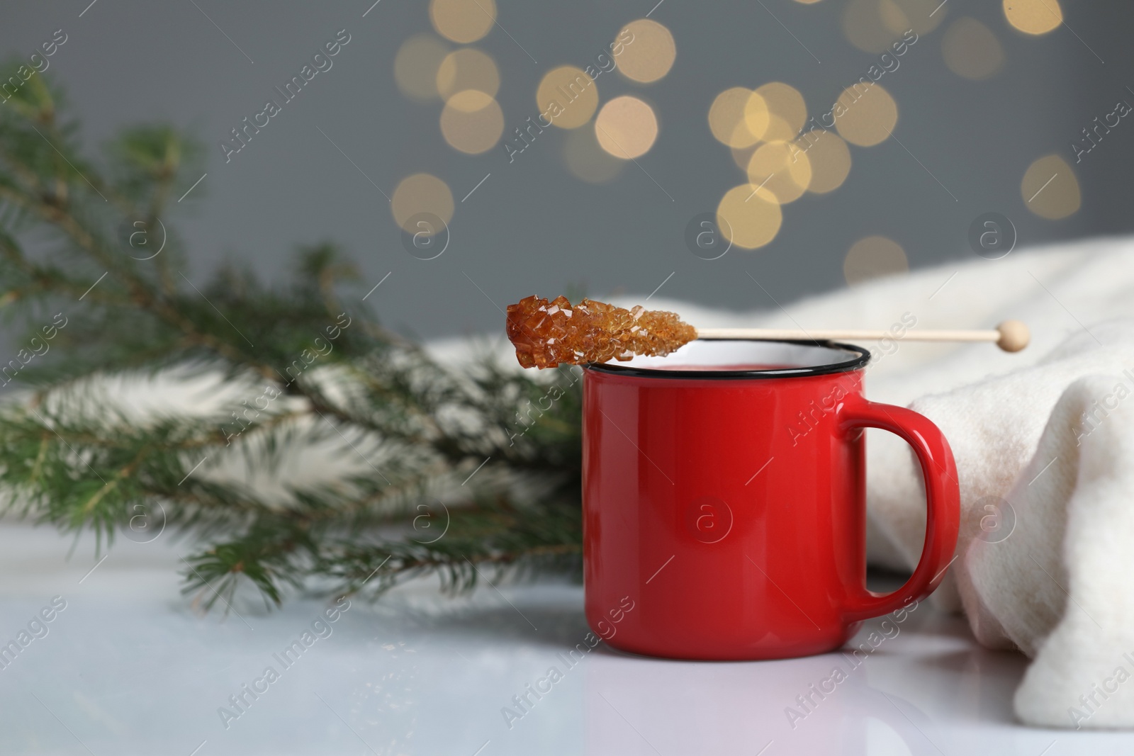 Photo of Stick with sugar crystals and cup of drink on white table against blurred festive lights, space for text