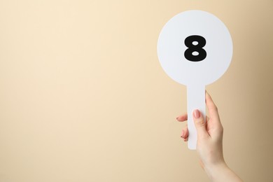 Photo of Woman holding auction paddle with number 8 on beige background, closeup. Space for text