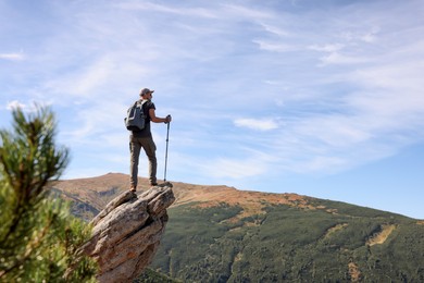 Photo of Man with backpack and trekking poles on rocky peak in mountains, back view