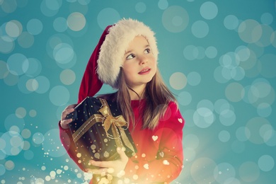Image of Cute child in Santa hat with Christmas gift on light blue background