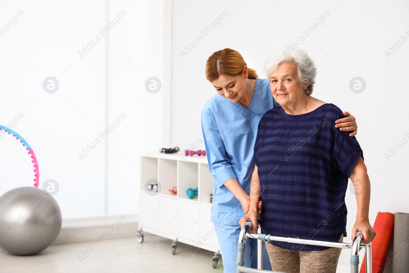 Photo of Caretaker helping elderly woman with walking frame indoors