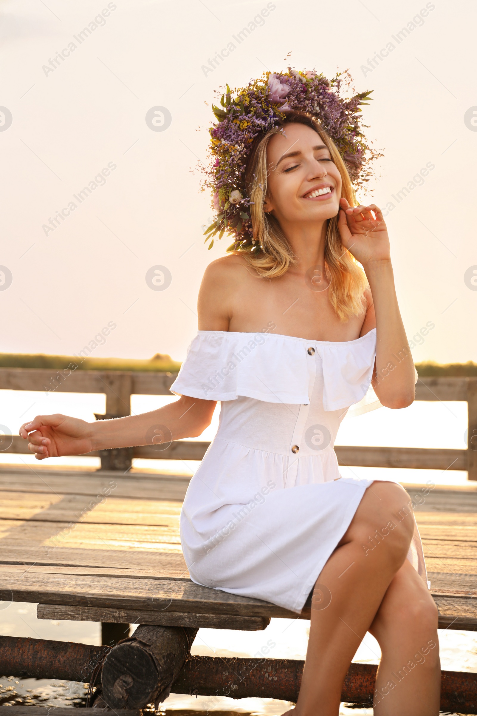 Photo of Young woman wearing wreath made of beautiful flowers on pier near river