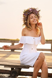 Photo of Young woman wearing wreath made of beautiful flowers on pier near river