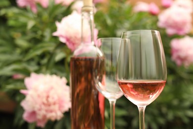 Photo of Glasses and bottle with rose wine against beautiful peonies, closeup. Space for text