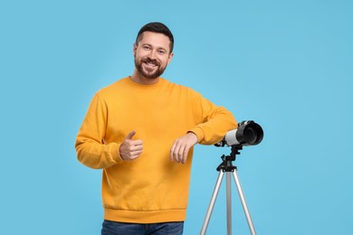 Smiling astronomer with telescope showing thumb up on light blue background