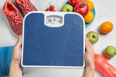 Photo of Woman holding scales over table with healthy food and sport equipment, top view. Weight loss