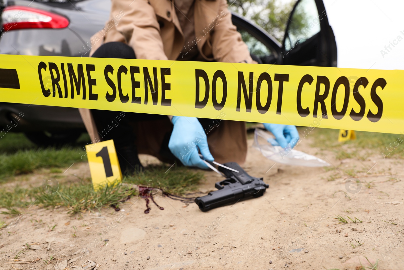 Photo of Investigator in protective gloves working with evidence at crime scene outdoors, focus on yellow tape
