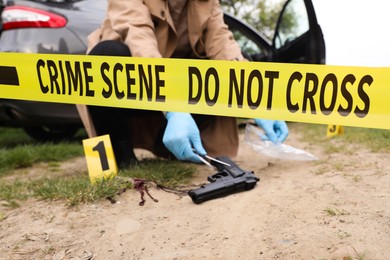 Investigator in protective gloves working with evidence at crime scene outdoors, focus on yellow tape