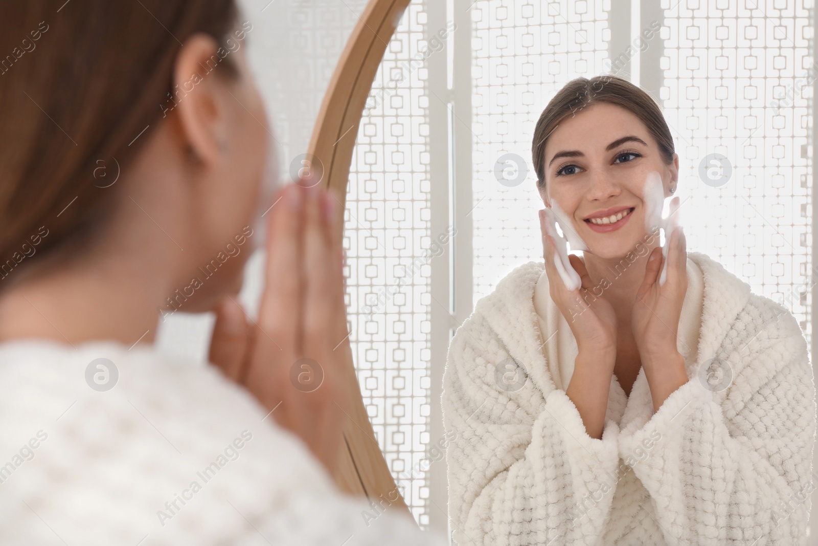 Photo of Young woman applying cleansing foam onto her face near mirror in bathroom