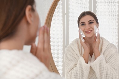 Photo of Young woman applying cleansing foam onto her face near mirror in bathroom