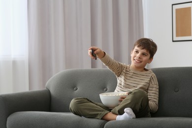 Photo of Little boy holding bowl of popcorn and changing TV channels with remote control on sofa at home